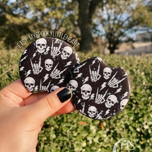 May include: Two black and white round buttons with a skull and lightning bolt pattern. The buttons feature a white skull with a white lightning bolt and a white skeleton hand making the rock and roll hand gesture. The text "ETSY: MYSPOOKYLITTLEHEART" is visible in the top left corner of the image.