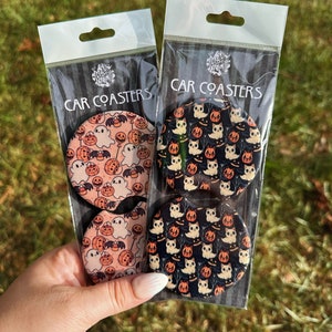May include: Two sets of Halloween-themed car coasters. Each set includes four coasters with different designs featuring ghosts, pumpkins, and cats. The coasters are round and have a white background with colorful designs.