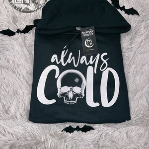 May include: Black hooded sweatshirt with a white skull and the text "always cold" printed on the front. The sweatshirt has a small tag attached to the drawstring with the text "Handmade with love" and a logo.