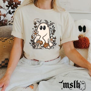 May include: Cream-colored t-shirt featuring a cartoon ghost with large eyes, surrounded by flowers, mushrooms, and bats. The shirt has short sleeves and a relaxed fit. The text "mslh" and a ghost graphic are on the sweatpants.