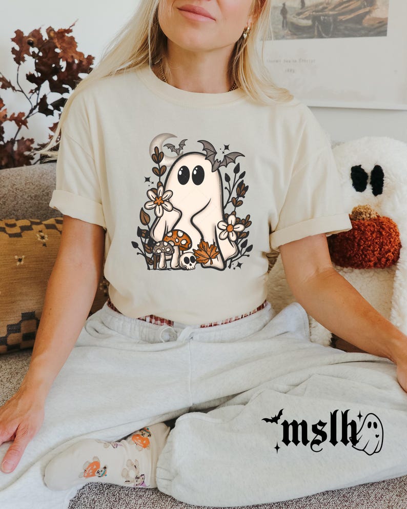 May include: Cream-colored t-shirt featuring a cartoon ghost with large eyes, surrounded by flowers, mushrooms, and bats. The shirt has short sleeves and a relaxed fit. The text "mslh" and a ghost graphic are on the sweatpants.