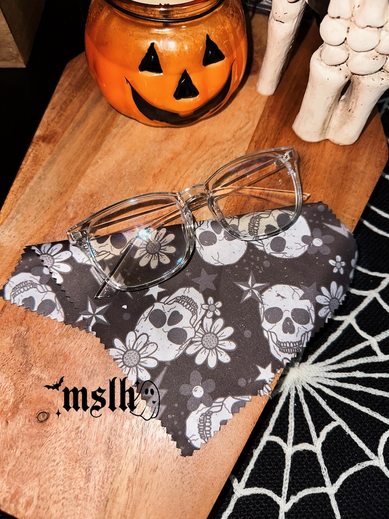 May include: A clear pair of glasses on a black microfiber cleaning cloth with a skull and flower pattern. The cloth is on a wooden surface with a jack-o-lantern and a skeleton hand in the background. The text "mslh" is printed on the cloth.