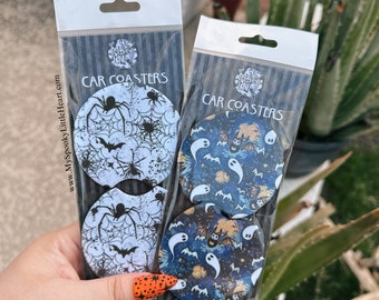 May include: Two sets of Halloween-themed car coasters. One set features black spiders and bats on a white web background. The other set features white ghosts and bats on a dark blue starry night background. Both sets are packaged in clear plastic bags with black and white striped backing. The text "CAR COASTERS" is printed on the bags.