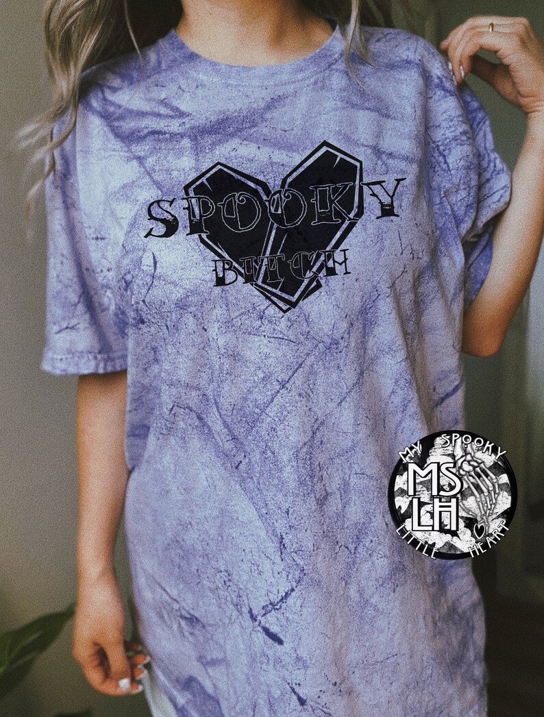May include: A purple tie-dye t-shirt with the text "Spooky Bitch" in black lettering. The text is inside a heart shape made of two black coffin shapes. The shirt also has a small black and white logo on the bottom right corner that says "My Spooky Little Heart".