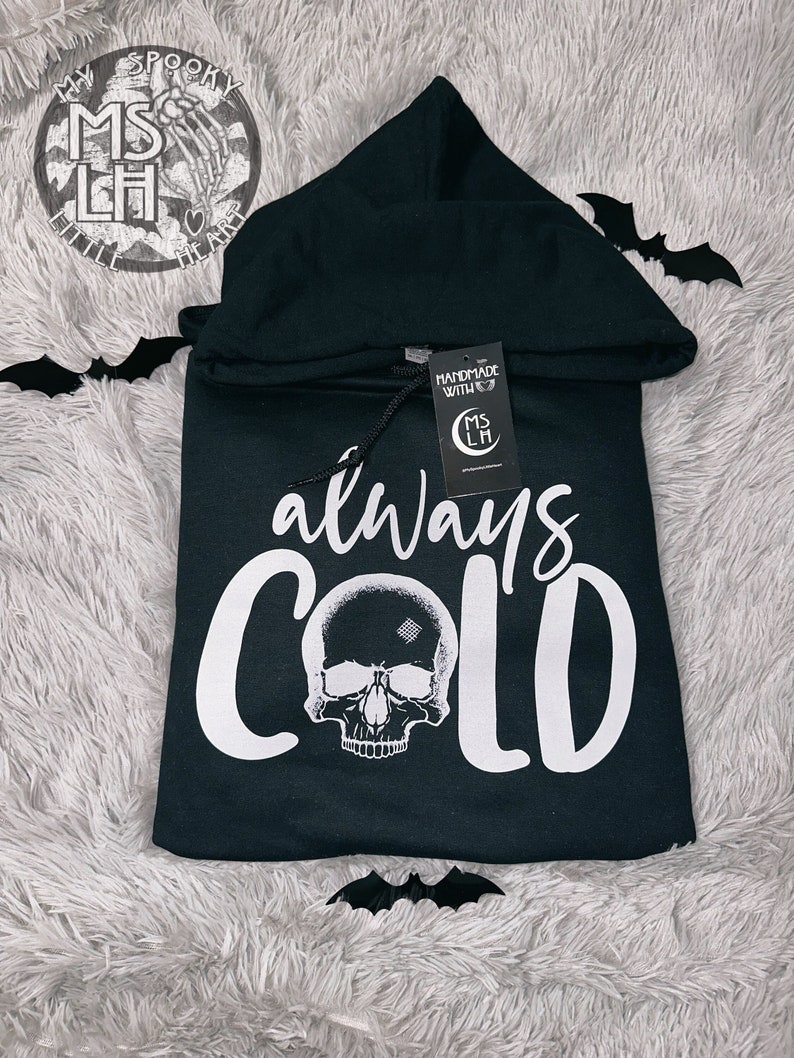 May include: Black hooded sweatshirt with a white skull and the text "always cold" printed on the front. The sweatshirt has a small tag attached to the drawstring with the text "Handmade with love" and a logo.