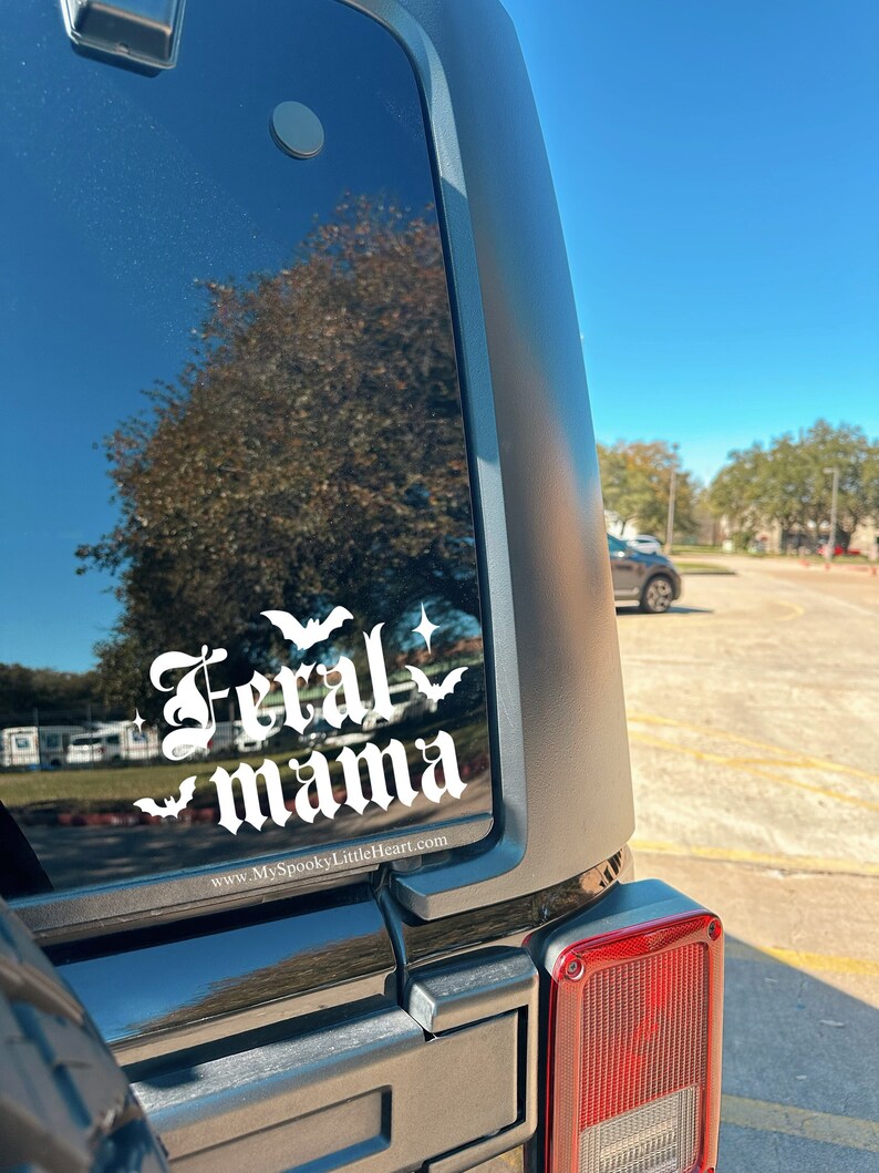 May include: A black Jeep with a white vinyl decal on the back window that reads "Feral mama" with bats and stars. The website www.MySpookyLittleHeart.com is printed below the decal.