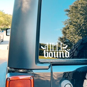 May include: A white vinyl decal with the words "Hell Bound" in a gothic font. Two bats are on either side of the word "Hell". The decal is on the back window of a black car.