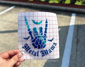 May include: A white vinyl sticker featuring a hand gesture with the words "Metal Mama" in a gothic font. The design includes bats and a gradient of blue and teal colors. The sticker is on a red grid background.