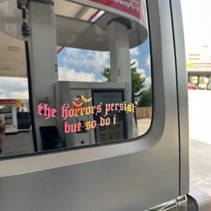 May include: A vinyl decal applied to a vehicle window featuring the text "the horrors persist but so do i" in a gothic-style font with a pink to orange gradient. Three small orange bat silhouettes are positioned above the text.