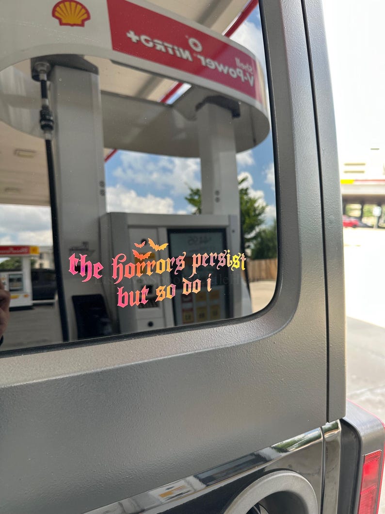 May include: A vinyl decal applied to a vehicle window featuring the text "the horrors persist but so do i" in a gothic-style font with a pink to orange gradient. Three small orange bat silhouettes are positioned above the text.