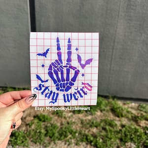 May include: A square decal featuring a purple and blue gradient skeleton hand making a peace sign, surrounded by small bat silhouettes and stars. The text "Stay weird" is printed in a gothic font below the hand, all on a white grid background.