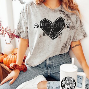May include: A woman wearing a gray t-shirt with the text "Spooky Bitch" in a black heart-shaped coffin design. She is wearing blue jeans and white sneakers. She is holding a white travel mug with a black design of a skeleton hand holding a skull with the text "My Spooky Bitch Life" around it.