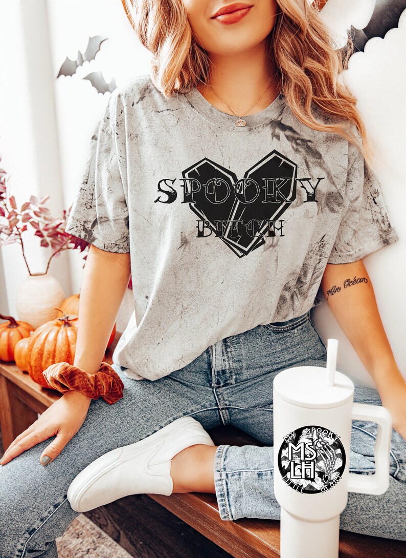 May include: A woman wearing a gray t-shirt with the text "Spooky Bitch" in a black heart-shaped coffin design. She is wearing blue jeans and white sneakers. She is holding a white travel mug with a black design of a skeleton hand holding a skull with the text "My Spooky Bitch Life" around it.