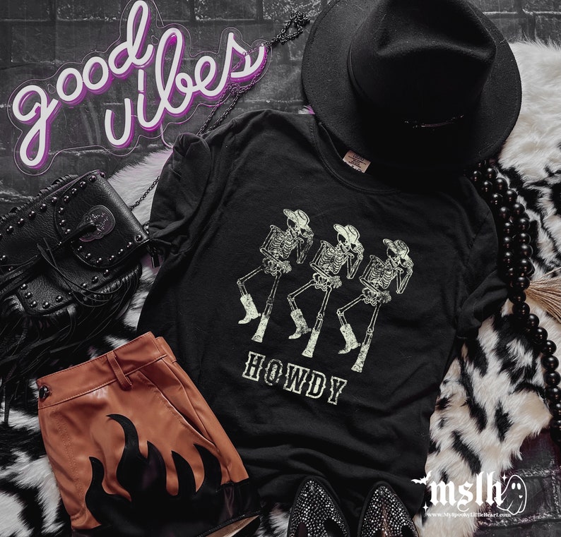 May include: Black t-shirt with a graphic of three dancing skeletons wearing cowboy hats and boots, with the word "HOWDY" below. The image includes a black hat, purse, and brown shorts with flame details, and the words "good vibes" in neon.