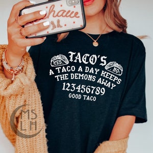 May include: A black t-shirt with a white graphic design that says "Taco's A Taco A Day Keeps The Demons Away 123456789 Good Taco". The graphic includes two stylized tacos with the words "Yes" and "No" on them.