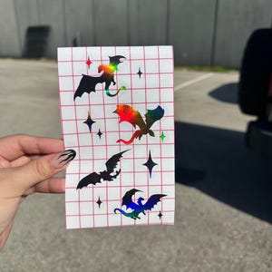 May include: A rectangular white sticker sheet featuring a red grid pattern and four distinct dragon silhouettes. The dragons display a vibrant holographic rainbow finish, accompanied by several small four-pointed star shapes scattered across the sheet.