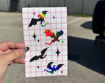 May include: A rectangular white sticker sheet featuring a red grid pattern and four distinct dragon silhouettes. The dragons display a vibrant holographic rainbow finish, accompanied by several small four-pointed star shapes scattered across the sheet.
