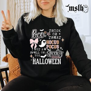 May include: Black Halloween-themed sweatshirt with the words "Boo," "Trick or Treat," "Hocus Pocus," and "Spooky" in a mix of fonts and colors. A pink bow and a ghost graphic are also featured.