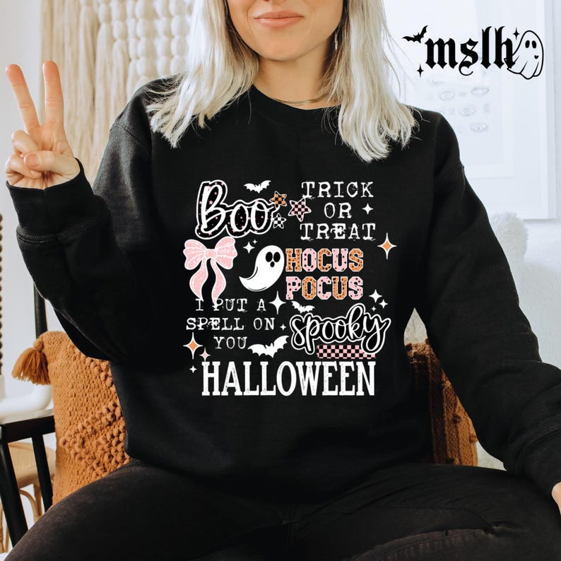 May include: Black Halloween-themed sweatshirt with the words "Boo," "Trick or Treat," "Hocus Pocus," and "Spooky" in a mix of fonts and colors. A pink bow and a ghost graphic are also featured.