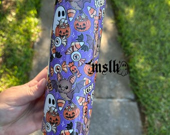 May include: A purple Halloween-themed tumbler featuring a repeating pattern of bats, ghosts, candy corn, and jack-o'-lanterns. The tumbler is cylindrical and appears to be made of stainless steel. The design is colorful and whimsical, perfect for the spooky season.