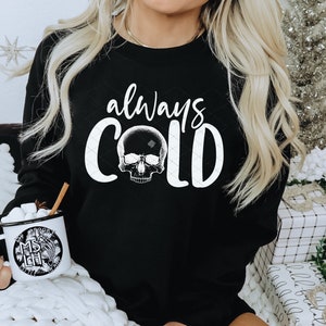 May include: A black sweatshirt with the text "always cold" in white lettering and a skull graphic. The sweatshirt is being worn by a person sitting on a white blanket.