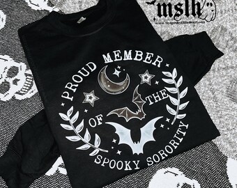May include: Black sweatshirt with a white graphic print that says "Proud Member of the Spooky Sorority". The graphic features a crescent moon, stars, and bats.