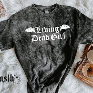 May include: Dark gray acid wash t-shirt with the text "Living Dead Girl" in white gothic lettering, with bat wing accents. The shirt is on a white fluffy surface, with a skull and candle in the background.