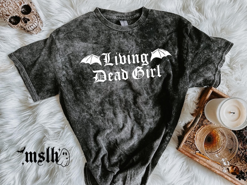 May include: Dark gray acid wash t-shirt with the text "Living Dead Girl" in white gothic lettering, with bat wing accents. The shirt is on a white fluffy surface, with a skull and candle in the background.