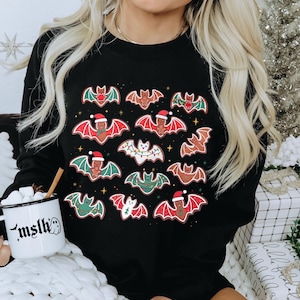 May include: Black sweatshirt featuring a pattern of gingerbread bats with red, green, and white accents. Some bats wear Santa hats or are decorated with Christmas lights. A white mug with the text "mslh" is held in the image.