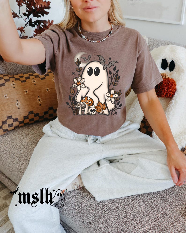 May include: A brown t-shirt featuring a cartoon ghost with floral and autumnal elements. The ghost has large black eyes and is surrounded by flowers, mushrooms, and leaves. The shirt has short sleeves and a relaxed fit.