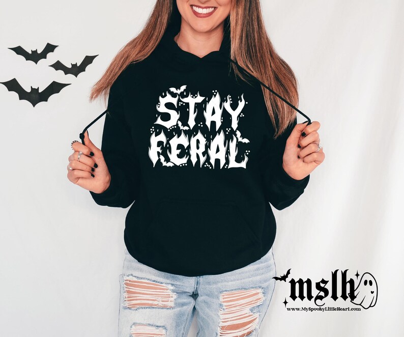 K&ouml;nnte beinhalten: Schwarzer Kapuzenpullover mit wei&szlig;em Text, der "Stay Feral" lautet, mit Flederm&auml;usen, die den Text umgeben. Das Model tr&auml;gt den Pullover mit zerrissenen Jeans.