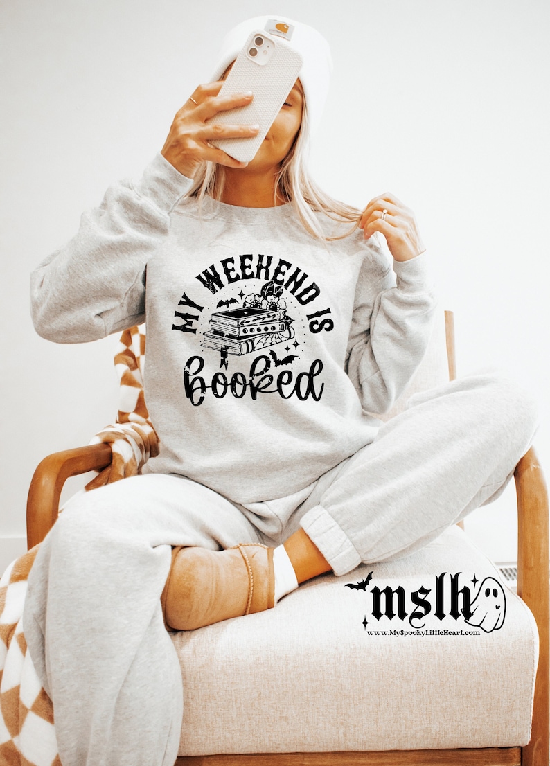 K&ouml;nnte beinhalten: Ein graues Sweatshirt mit einer schwarzen Grafik, auf der "My Weekend is Booked" steht, mit einem Stapel B&uuml;cher, Flederm&auml;usen und Sternen. Die Grafik ist in einem Kreis.