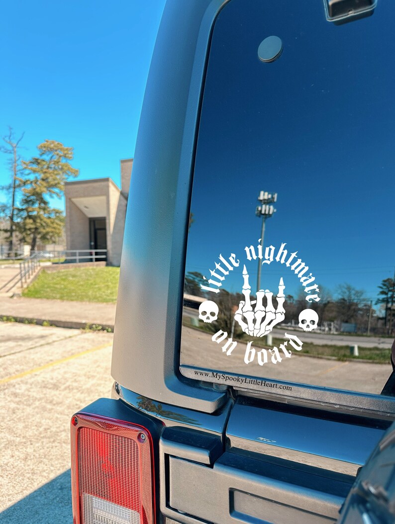 May include: A white vinyl sticker on a black car. The sticker says "little nightmare on board" in a gothic font with a skeleton hand giving the middle finger and two skulls on either side. The website www.myspookylittleheart.com is printed at the bottom.
