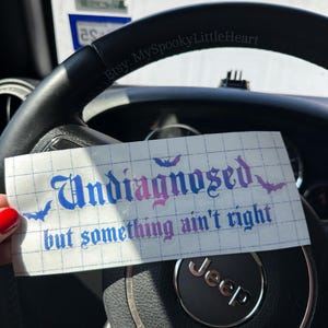 May include: A white sticker with the words "Undiagnosed but something ain't right" in blue and pink gothic font, with bat silhouettes. The sticker is held over a black steering wheel with a Jeep logo.