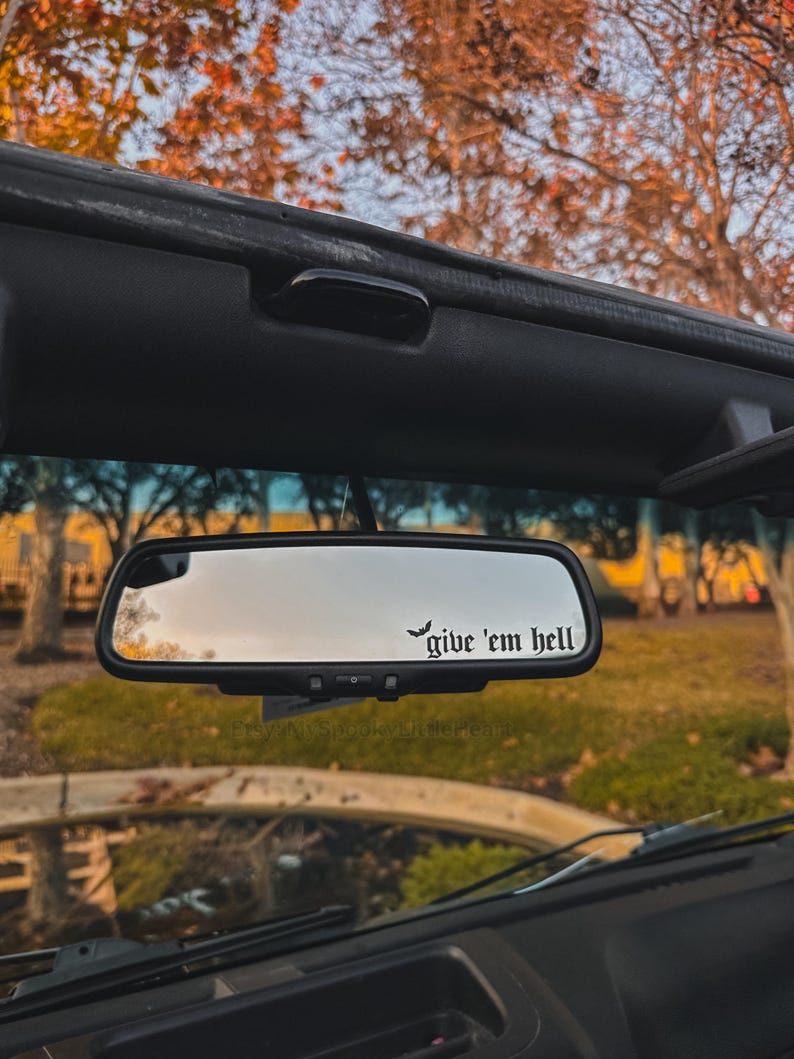 May include: A car's rearview mirror with the phrase "give 'em hell" in a gothic font. The mirror is rectangular and mounted inside a vehicle, reflecting a view of trees and a sunny day.