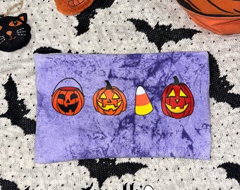 May include: A rectangular purple tie-dye fabric pouch featuring a graphic print of a jack-o'-lantern candy bucket, a jack-o'-lantern, a piece of candy corn, and another jack-o'-lantern. The text mslh with a ghost icon is printed below.