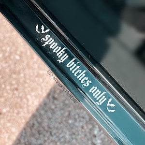 May include: A black and silver car door trim with a white vinyl decal that reads "Spooky Bitches Only" with bat wings on either side. The decal is on a black background and has a gothic font.
