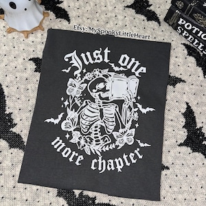 May include: Black t-shirt with a white graphic of a skeleton reading a book, surrounded by butterflies and foliage. The text reads "Just one more chapter." The Etsy shop name "MySpookyLittleHeart" is also visible.