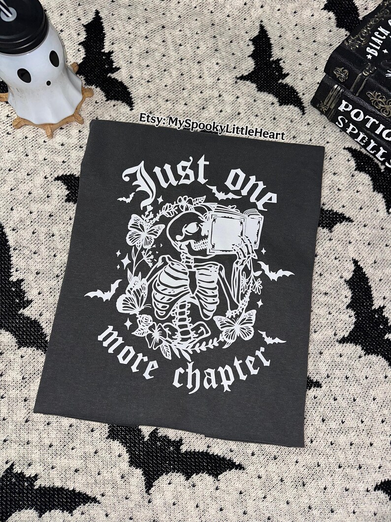 May include: Black t-shirt with a white graphic of a skeleton reading a book, surrounded by butterflies and foliage. The text reads "Just one more chapter." The Etsy shop name "MySpookyLittleHeart" is also visible.