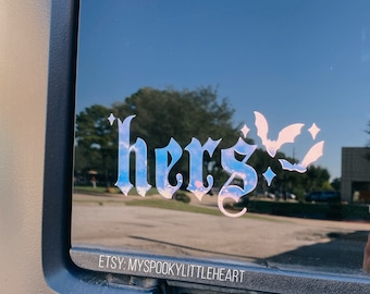 May include: A holographic sticker with the word "here" in gothic font and a bat silhouette. The sticker is on the back window of a black car. The text "ETSY: MYSPOOKYLITTLEHEART" is visible below the sticker.