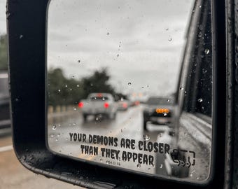 May include: A black rectangular vehicle side mirror with raindrops on the glass surface. The mirror features the printed text 'YOUR DEMONS ARE CLOSER THAN THEY APPEAR' in a bold, dark font, reflecting a road with blurred cars on a rainy day.