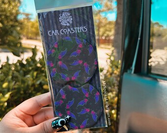 May include: Two round car coasters with a black background and a pattern of purple bats with pink accents. The coasters are packaged in a clear plastic bag with a white label that says "CAR COASTERS" and a website address.