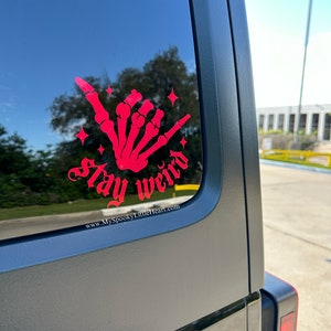 May include: A red skeleton hand making a shaka sign with the text "Stay Weird" and the website "www.MySpookyLittleHeart.com" below it. The sticker is on the back window of a black car.