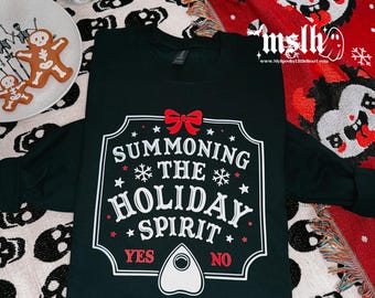 May include: Black long-sleeved shirt with a white graphic of a Ouija board with the text "Summoning the Holiday Spirit" and "Yes" and "No" options. The shirt is on a white and black skull patterned blanket.