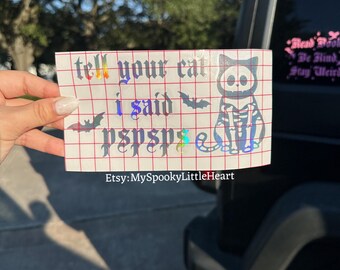 May include: A holographic vinyl decal featuring the text "tell your cat i said pspsps" in a gothic font, accompanied by two small bat silhouettes and a graphic of a cat skeleton. The design is printed on a white grid transfer paper.