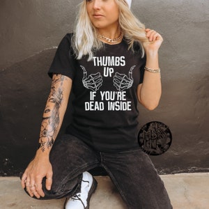 Pu&ograve; includere: Maglietta nera con una grafica bianca di due mani di scheletro che fanno un pollice in su con il testo "Thumbs Up If You're Dead Inside".