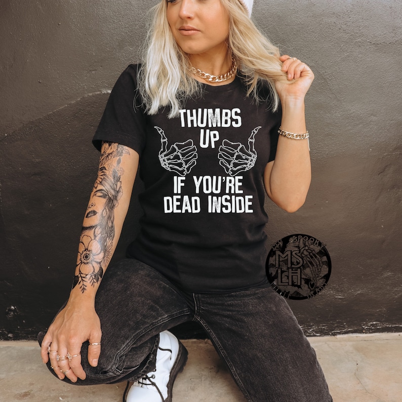 Pu&ograve; includere: Maglietta nera con una grafica bianca di due mani di scheletro che fanno un pollice in su con il testo "Thumbs Up If You're Dead Inside".