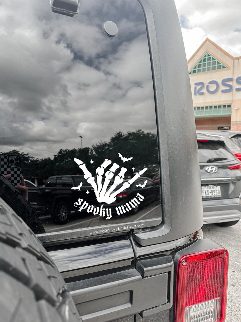 May include: A black car with a white vinyl decal on the back window. The decal features a skeleton hand making a shaka sign with the words "spooky mama" and a website address below.