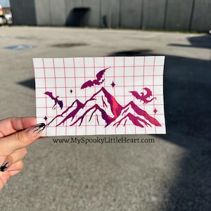 May include: A magenta holographic vinyl decal featuring a mountain range silhouette with three flying dragons and small four-pointed stars. The design is printed on a white grid transfer paper with the text www.MySpookyLittleHeart.com at the bottom.