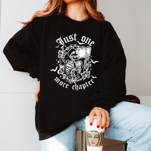 May include: Black sweatshirt with a white graphic of a skeleton reading a book, surrounded by flowers and bats. The text "Just one more chapter" is printed below the graphic. The sweatshirt is a casual style.